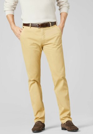 Man draagt beige chino's, een witte textuurtrui, een bruine leren riem en bruine suède loafers, staat met handen gedeeltelijk in de zakken.