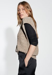 Gilet in maglia beige con finitura testurizzata, dotato di ampie aperture per le braccia e una vestibilità rilassata, abbinato a pantaloni neri a vita alta.