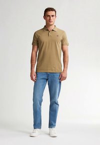 Khakifarbenes Poloshirt mit kurzen Ärmeln, Kragen und Logo auf der Brust, kombiniert mit hellblauen Jeans und weißen Turnschuhen.