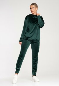 Aksamitny dres w ciemnozielonym kolorze o luźnym kroju. Posiada wysoką kołnierzową stójkę i długie rękawy, zestawiony z dżinsami zwężającymi się ku dołowi oraz białymi sneakersami.