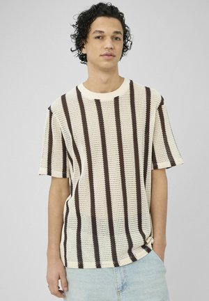 Jeune homme aux cheveux bouclés portant une chemise en tricot crème à manches courtes avec des rayures verticales marron et un jean bleu clair, sur un fond uni.