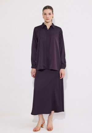 Γυναίκα στέκεται φορώντας μια χαλαρή σκούρα μωβ μπλούζα με μακριά μανίκια και αντιστοιχιστή μακριά φούστα με ροδακινί σανδάλια με χαμηλό τακούνι μπροστά σε λευκό τοίχο.