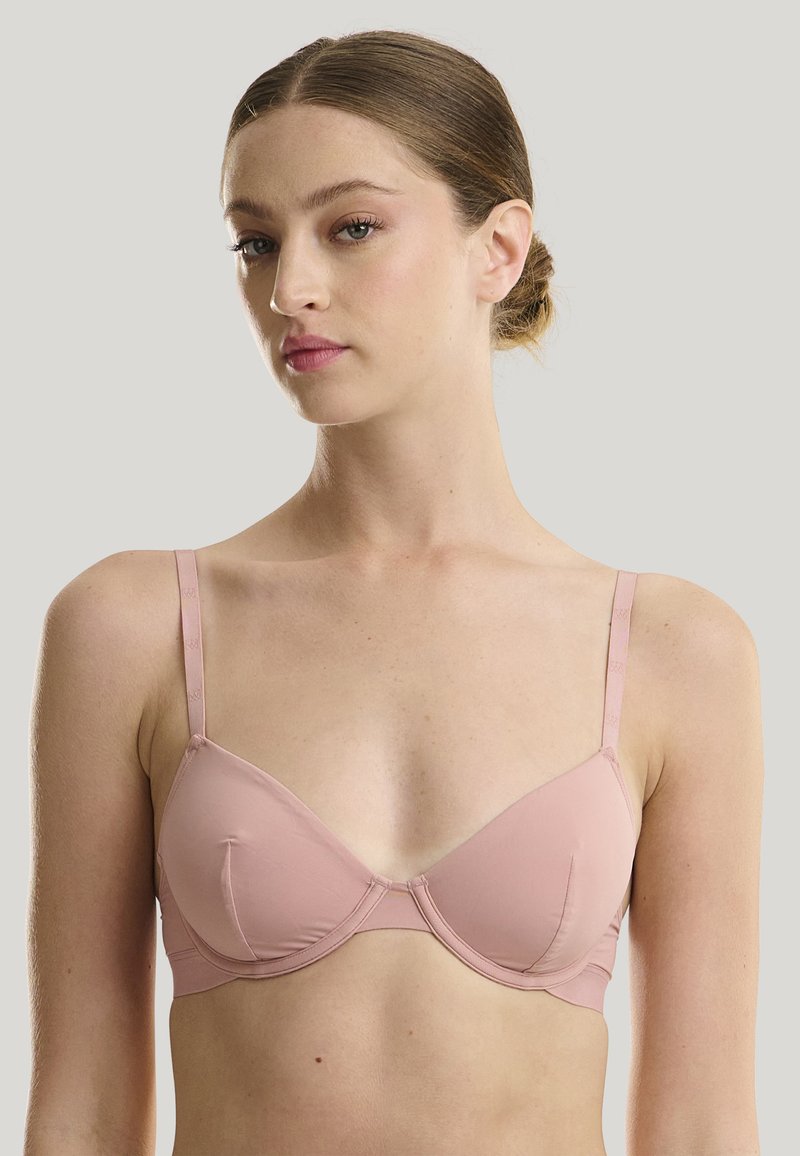 Soutien-gorge rose clair avec des bonnets moulés et des bretelles ajustables. Présente une texture lisse et des détails de couture délicats le long des bords.