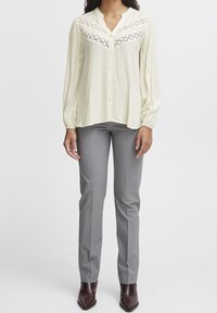 Blouse crème clair avec des détails en dentelle au niveau de la poitrine ; manches longues. Associée à un pantalon gris à rayures et des bottines marron.