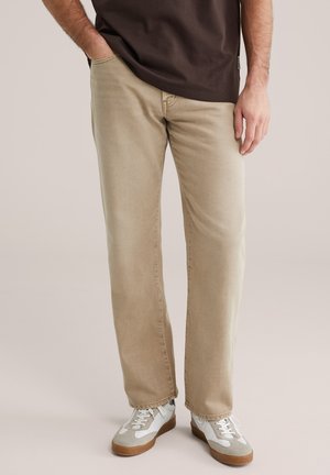 Pantalon beige à jambes droites en tissu mélangé coton, comprenant un design à cinq poches. Associé à des baskets grises et blanches.
