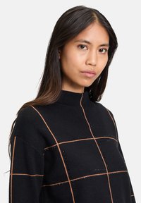 Schwarzer Pullover mit hohem Kragen, verziert mit einem Karomuster aus beigen und schwarzen Strasssteinen, aus weichem Strickmaterial gefertigt.