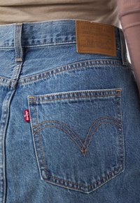 Vue arrière d'un jean en denim bleu Levi's avec un patch en cuir de marque et une étiquette rouge Levi's sur la poche arrière, porté avec un haut beige.