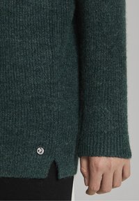 Pull en maille de couleur vert foncé avec une texture côtelée, fente sur le côté, et un petit accent en logo argenté au bas de l'ourlet. Matière douce et chaude.