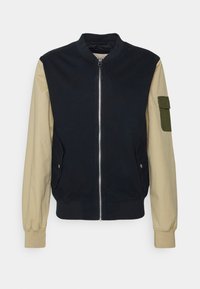 Bomberjacka i bomull med en marinblå kropp, beige ärmar och en grön ficka på vänster arm. Den har dragkedja fram och sidofickor.