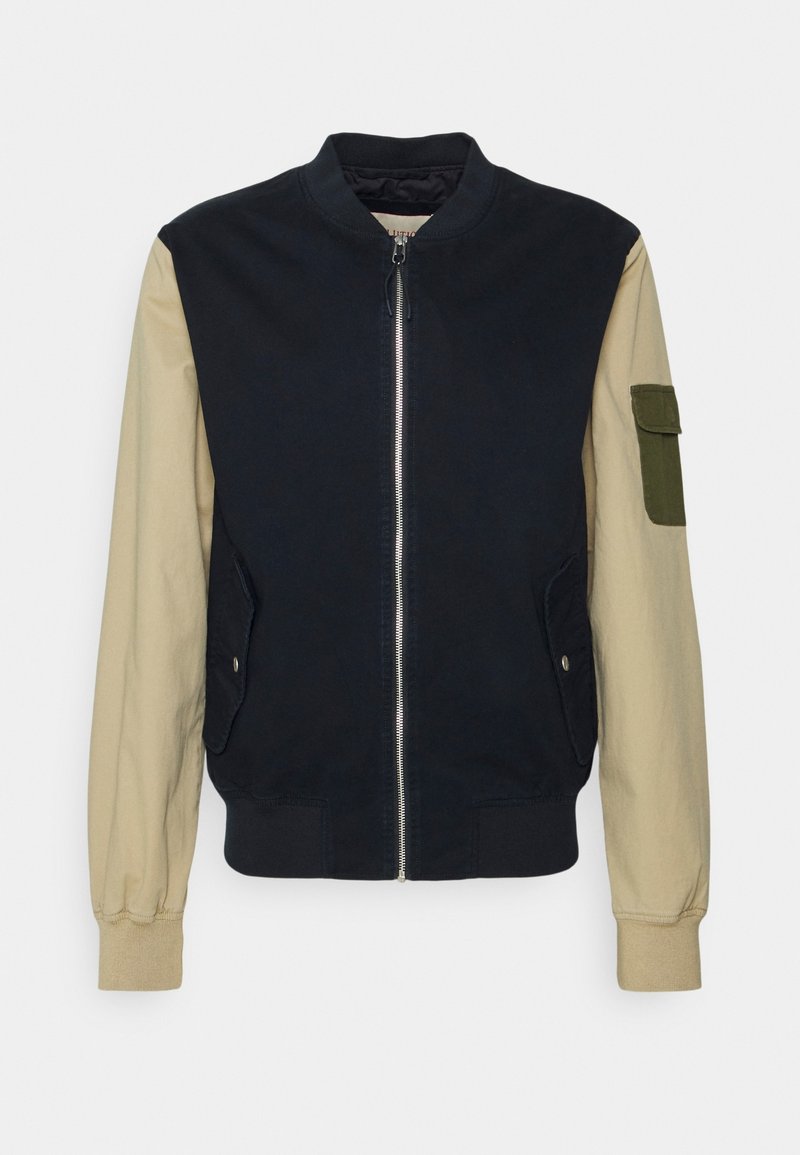 Bomberjacka i bomull med en marinblå kropp, beige ärmar och en grön ficka på vänster arm. Den har dragkedja fram och sidofickor.