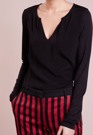 Person iført en sort langærmet V-hals top og højtaljede røde bukser med sorte lodrette striber, hænder i lommerne.