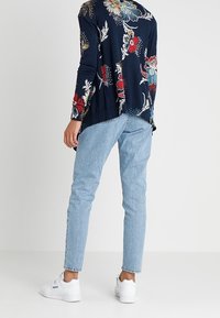 Marinblus med lång ärm i blommönster med livliga röda och blå mönster, ihop med ljusblå denimjeans och vita sneakers.