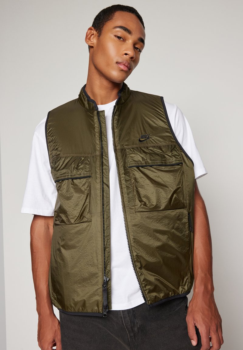 Gilet sans manches vert olive avec fermeture éclair sur le devant, quatre poches et texture cousue. Présente des accents noirs et un petit logo sur la poitrine.