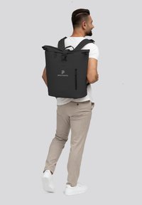 Mochila impermeable negra con diseño de parte superior enrollable, correas de hombro ajustables y un bolsillo con cremallera lateral. Logo mostrado en la parte frontal.