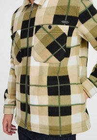 Veste en polaire à motifs carreaux beige, vert et noir. Dispose de deux poches poitrine avec des boutons noirs et d'une fermeture éclair à l'avant. Texture douce.