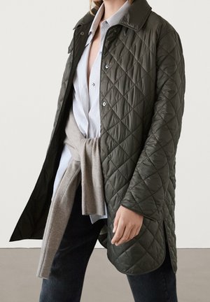 Femme portant un manteau matelassé vert foncé sur une chemise rayée avec un pull beige noué à la taille et un pantalon foncé, debout devant un fond uni.