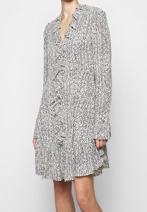 Biała sukienka z długim rękawem z czarnym nieregularnym wzorem w kropki, z falbankami na przodzie i mankietach, noszona przez stojącą osobę z luźno opuszczonymi rękami.