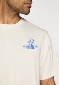 Μπεζ βαμβακερό t-shirt με μπλε γραφικό ενός σκιέρ και το λογότυπο "THE NORTH FACE" στο αριστερό στήθος. Λεία υφή, στρογγυλή λαιμόκοψη.