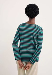 Žmogus atsuko nugarą, vilkintis ilgomis rankovėmis marškiniu su horizontaliomis melsvai žalia, pilka, juoda juostelėmis ir šviesiomis kelnėmis, rankas suspaudęs už nugaros.