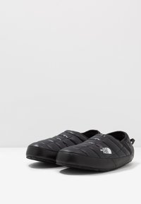 Black quilted slip-on shoes with a smooth exterior, featuring a rubber sole and a white logo on the side. Cushioned for comfort.