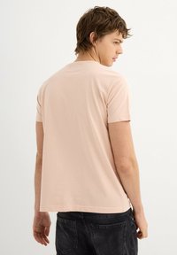 Camiseta de manga corta rosa claro hecha de algodón suave, con un cuello redondo clásico y un corte relajado, combinada con jeans gris oscuro.