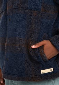 Marineblå sherpa-jakke med brune ternede detaljer, knaplukning, lommehuller og et mærke med "EST. 1992" på kanten. Blødt, tekstureret stof.