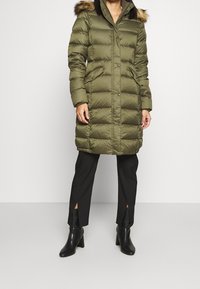 Cappotto imbottito verde oliva con cappuccio foderato in pelliccia, caratterizzato da un design trapuntato, bottoni a pressione e tasche laterali. Indossato sopra pantaloni neri su misura.