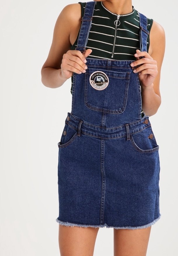 Denimoverallklänning i mörkblått med fransad nederkant, med justerbara axelremmar, en bröstficka och en rund patch på bröstet.