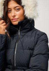 Sort pufferjakke med høj krave, featuring en aftagelig faux-fur hætte, quiltet tekstur og en sølvfarvet lynlås lukning.