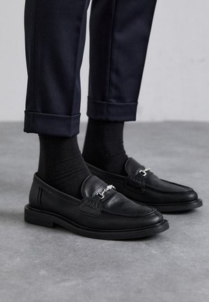 Moccasines de cuero negro con un acabado texturizado, detalle de herrajes plateados, combinados con calcetines oscuros y pantalones de corte navy con dobladillo.