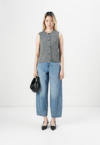 Mujer de pie frente a un fondo blanco, vistiendo una blusa sin mangas gris y abotonada, unos vaqueros de pierna ancha azules, unos zapatos negros de punta y sosteniendo un pequeño bolso negro.