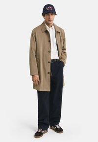 Cappotto lungo beige con bottoni, indossato su una camicia bianca, abbinato a pantaloni blu navy e sneaker marroni con accenti chiari. Berretto navy con logo "GANT".