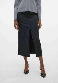 Jupe midi en cuir noir avec une fente à l'avant, texture lisse et design sur mesure. Associée à des talons aiguilles noirs et un haut gris clair.