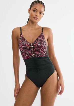 Kobieta ubrana w jednoczęściowy kostium kąpielowy z różowo-czarnym topem w zebra print i jednolitym czarnym marszczonym dołem, stojąca na jednolitym białym tle.