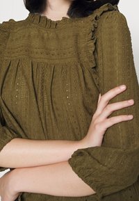 Olivgrüne Bluse aus strukturiertem Stoff, mit Rüschenausschnitt und Dreiviertelärmeln. Hat filigrane Muster und subtile Lochdetails.