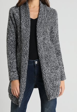 Vrouw draagt een zwart-wit gemêleerde, openvallende cardigan over een zwart V-hals top en donkerblauwe spijkerbroek.