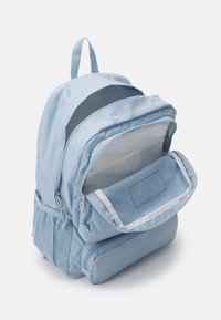 Mochila azul clara con múltiples compartimentos con cremallera, incluyendo un bolsillo frontal con solapa abierta y un bolsillo lateral de malla, sobre un fondo blanco.