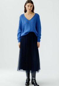 Pull en tricot bleu avec un col en V, associé à une jupe en tulle noir plissée, porté avec des bottes noires au genou. Matériaux lisses et texturés.