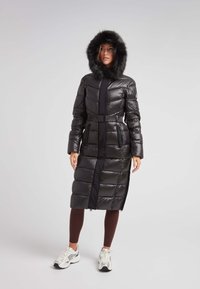 Abrigo de plumas negro con capucha forrada de piel, cintura ceñida con cinturón y diseño de acolchado vertical. Cuenta con bolsillos laterales y un corte hasta las rodillas.
