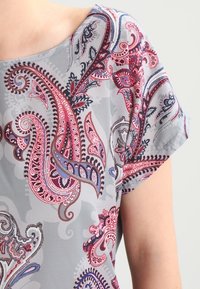 Kortärmad blus i ljusgrått tyg med ett färgglatt paisleymönster i nyanser av rosa, blått och mörkrött. Slät textur.