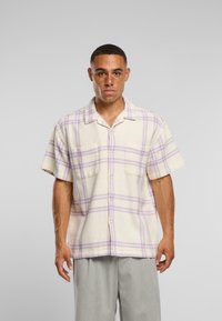 Camicia a maniche corte a quadri in crema con strisce lavanda, con due tasche sul petto e una vestibilità rilassata. Il tessuto appare testurizzato e intrecciato.