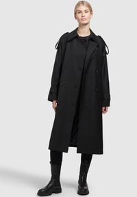 Černý trenčkot s dvouřadým zapínáním na knoflíky, širokými klopami a popruhy na ramenech. Nenošený přes černý outfit a spárovaný s černými bojovými botami.