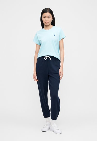 Gaiši zila kokvilnas T-krekls ar īsām piedurknēm un tumši zilām auklām viduklī, kopā ar tumšām sporta biksēm un baltām sporta kurpēm.