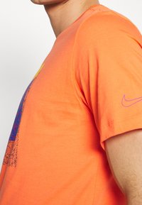 Muž oblečený v jasně oranžovém tričku Nike s fialovým logem na rukávu a pestrým grafickým vzorem na přední straně, zobrazený od ramene po hrudník.