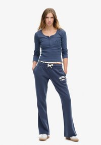 Blå ribbet langærmet top med knaplukning og logo, parret med blå sweatpants med et hvidt grafisk design og snørebånd i taljen.