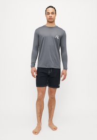Vīriešu pelēks garo piedurkņu rash guards ar logotipu, apvienots ar melniem peldbikšiem ar baltām šuvēm un sānu kabatām. Stāvoklis basām kājām.
