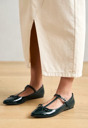 Green patent leather ballet flats with a T-strap, bow detail on the toe, and low wedge heel, paired with a cream colored ankle-length skirt.
