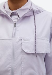 Κοντινό πλάνο ενός ατόμου που φοράει ένα ελαφρώς μωβ μπουφάν Columbia με φερμουάρ μπροστά, τσέπη στο στήθος, γιακά με κουμπιά και κουκούλα με κορδόνι.