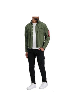 Olive green, button-up shirt with chest pockets and a red patch on the sleeve, paired with black cargo pants and white sneakers.