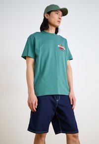 Camiseta de algodón verde con mangas cortas y un gráfico colorido en el pecho, combinada con pantalones cortos de mezclilla azul marino con costuras blancas.